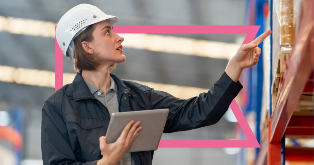 White woman in a white hard hat and blue smock holding an ipad in front of inventory shelves