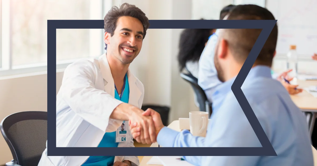 A smiling healthcare professional in a white coat and blue scrubs shakes hands with a business professional across a table.