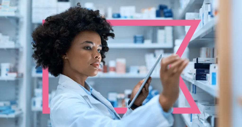 This image shows a woman in a pharmacy setting, wearing a white lab coat, suggesting she is a pharmacist or healthcare professional. She is standing in front of shelves filled with various medications, attentively selecting a box from the shelf. She holds a tablet in one hand, possibly using it to manage inventory or access patient records. The background consists of neatly arranged medication boxes, creating a clean and organized environment. The image has a modern, professional tone, with a pink geometric shape framing part of the image for added visual interest.