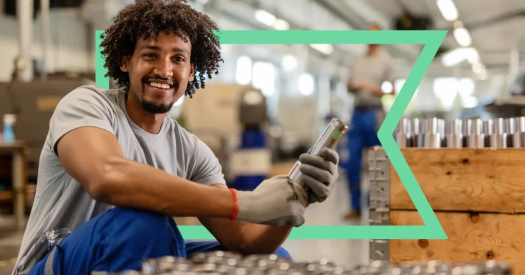 Smiling worker holding a tool in a manufacturing setting, seated in front of machinery and metal components, with a green geometric frame in the background.