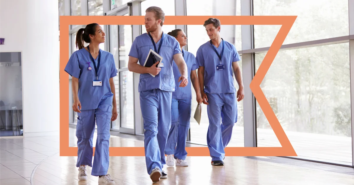 Four healthcare workers in blue scrubs walking through a wide hospital hallway